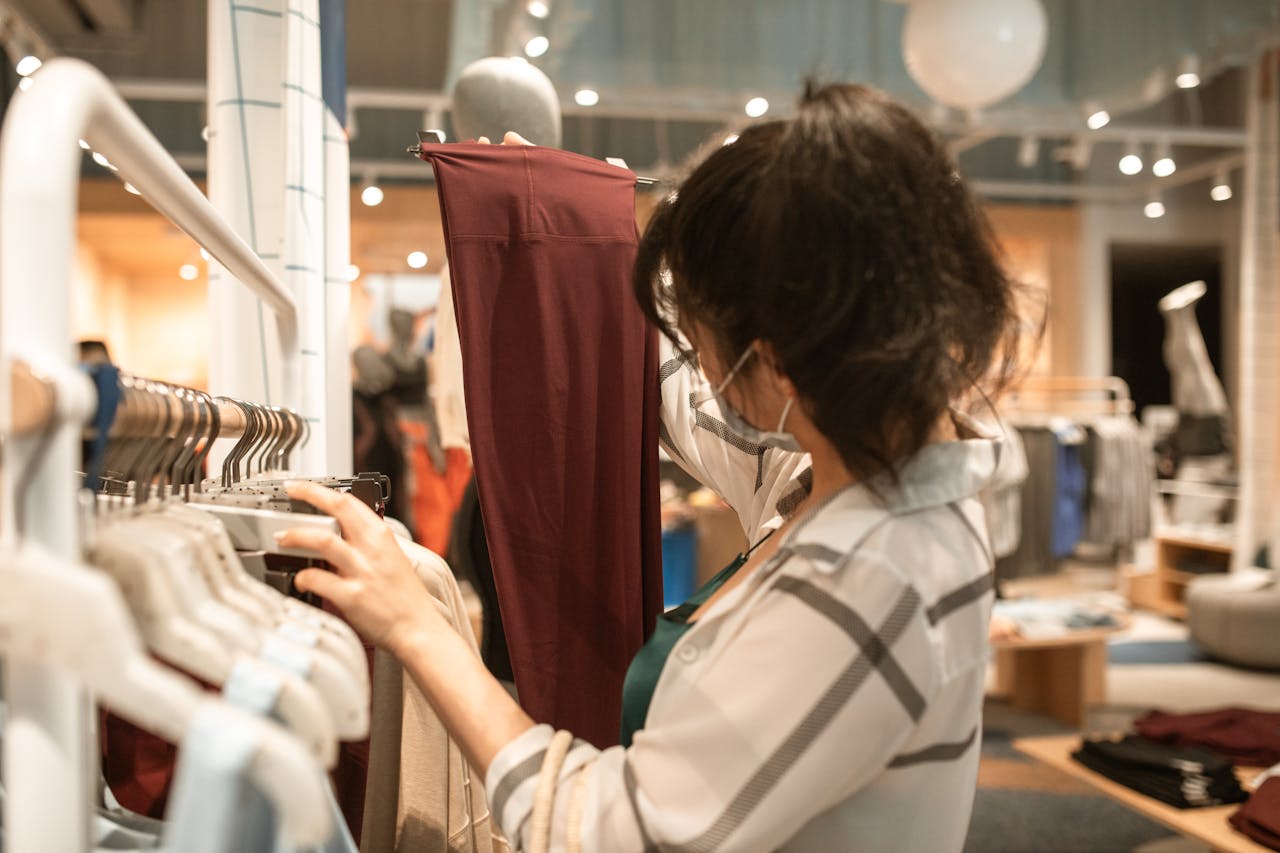 Woman wearing mask in a store shopping for clothes, selecting burgundy pants.